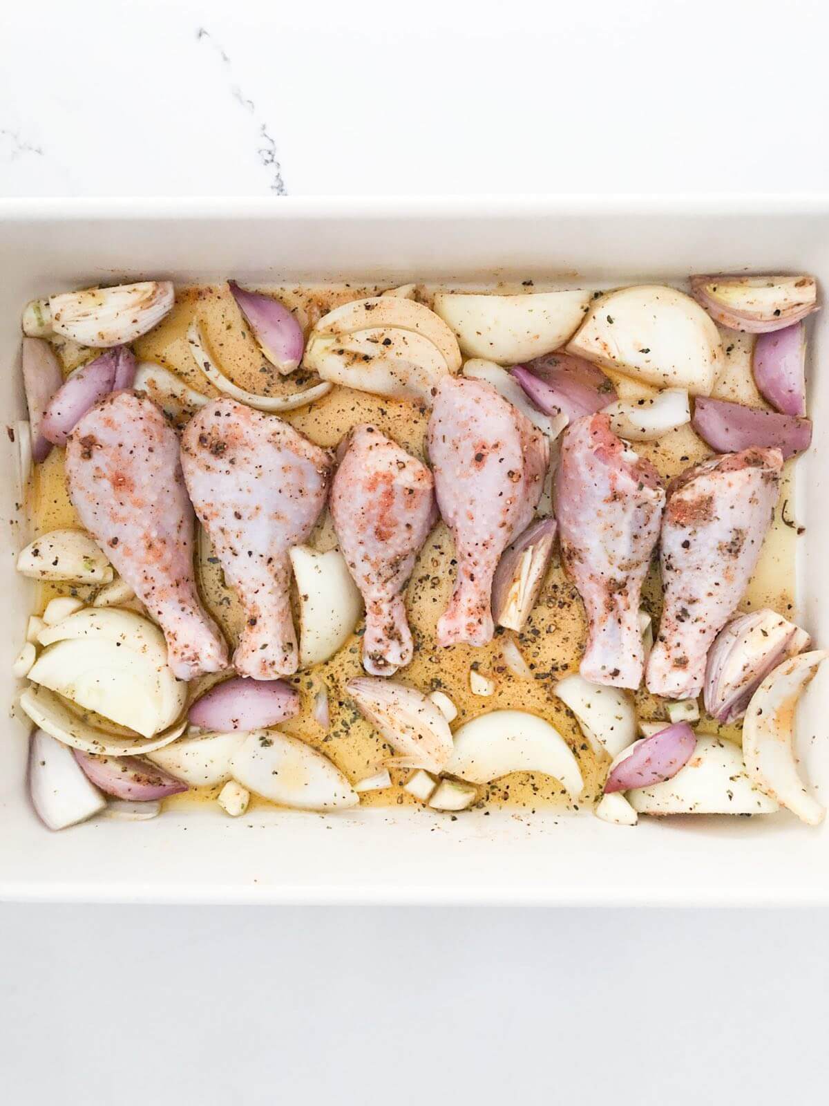 uncooked chicken drumsticks in olive oil, onions, garlic, and seasonings in a baking dish.