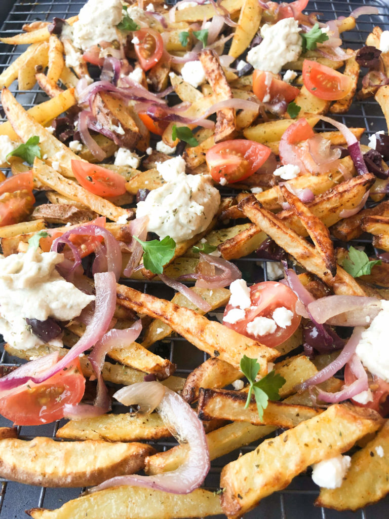 homemade greek fries topped with feta, olives, tomatoes, and red onions on a baking sheet.