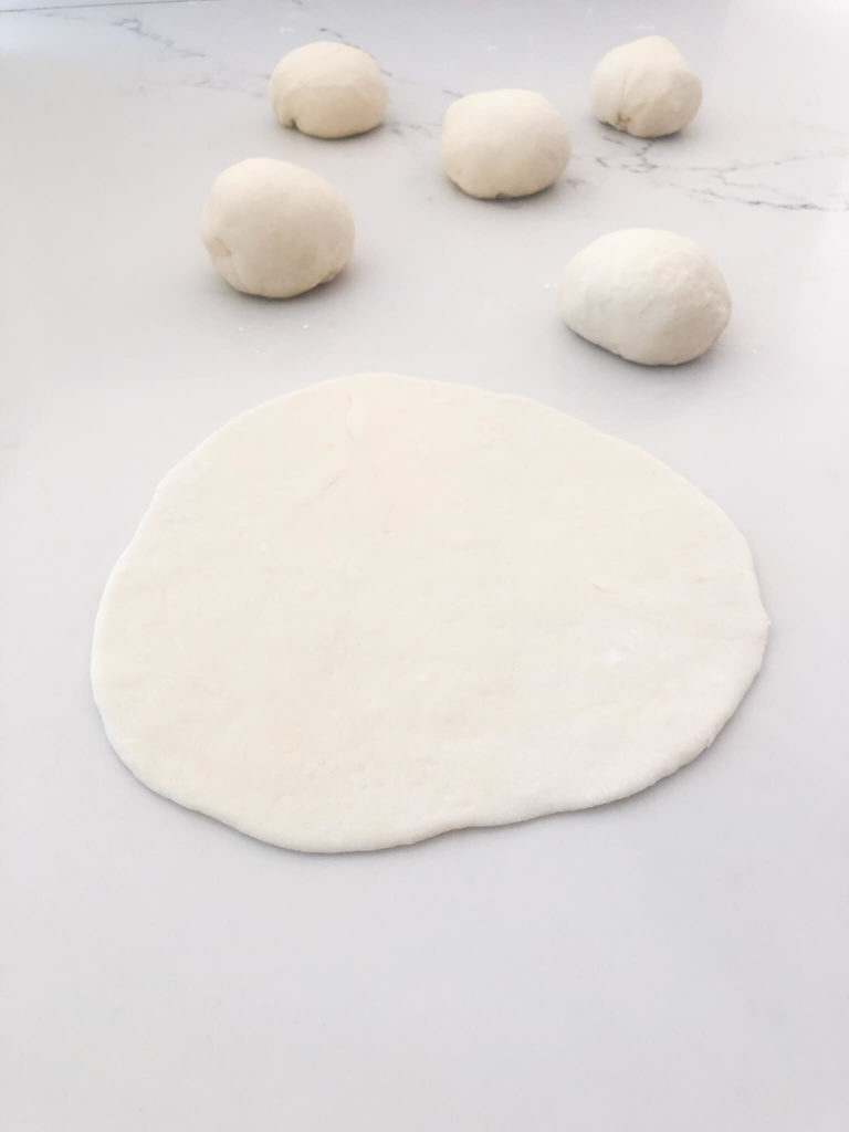 pita bread dough rolled out into a circle with a few dough balls behind it on a kitchen counter.
