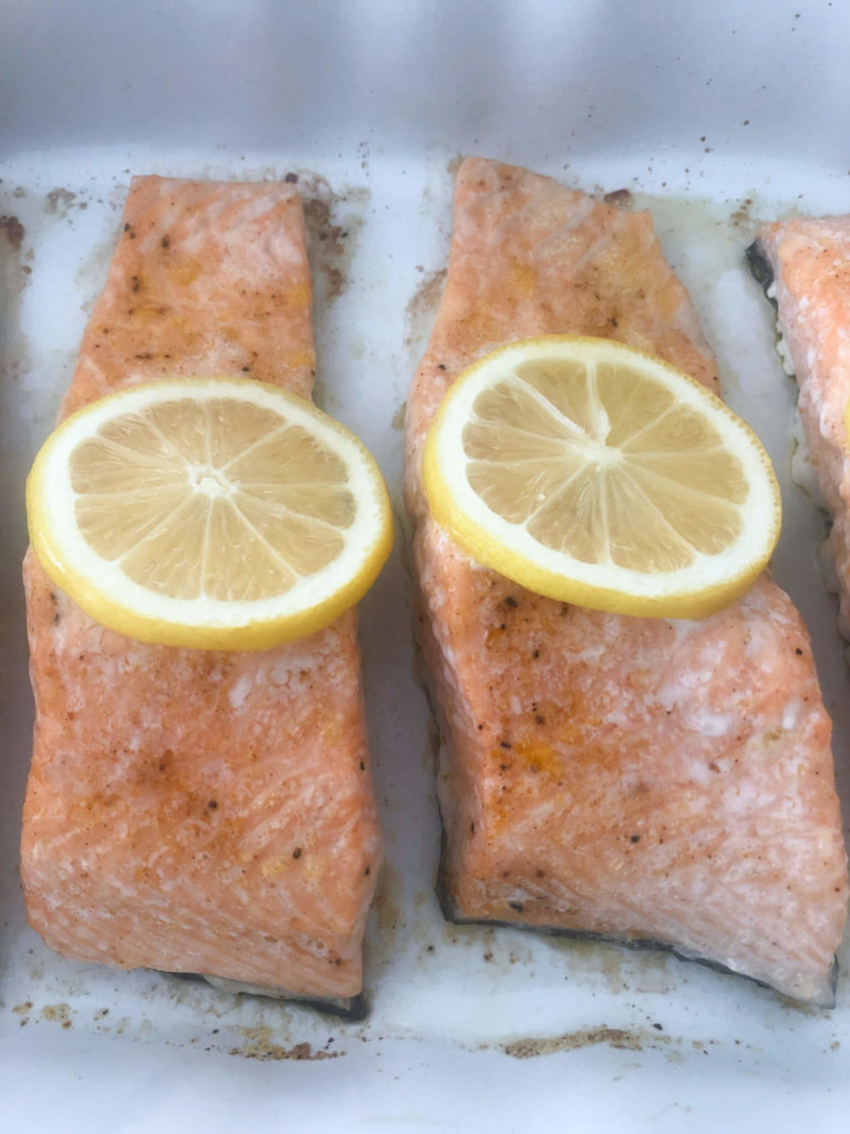 two pieces of baked salmon with a lemon slice in a white baking dish.