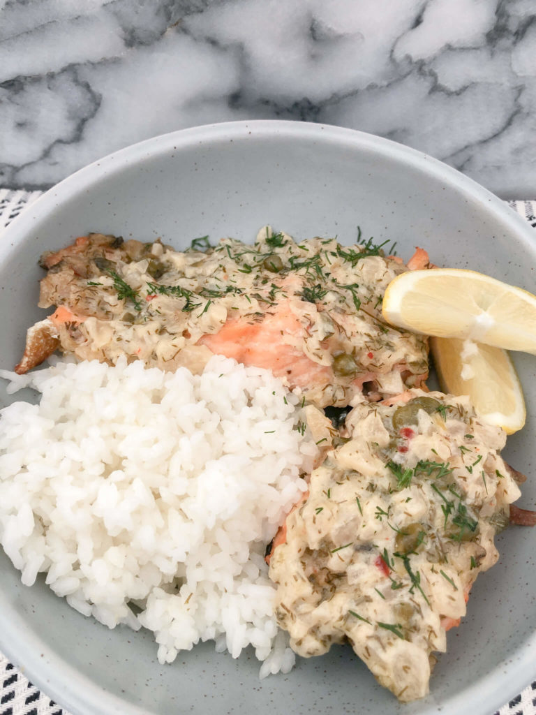two pieces of salmon with creamy dill mayo sauce, white rice and two lemon wedges in a light blue bowl.