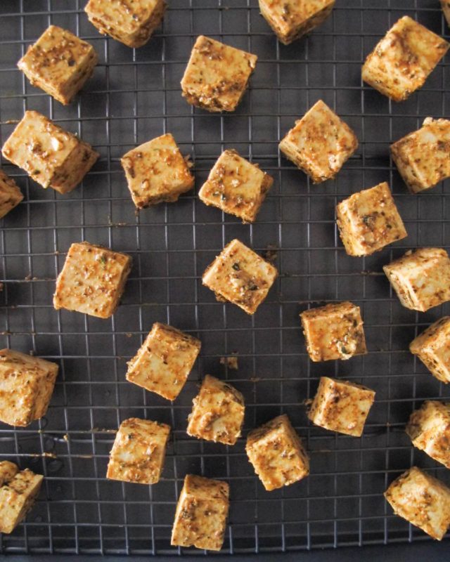 Cubes of Greek marinated tofu spread out on a black baking sheet.