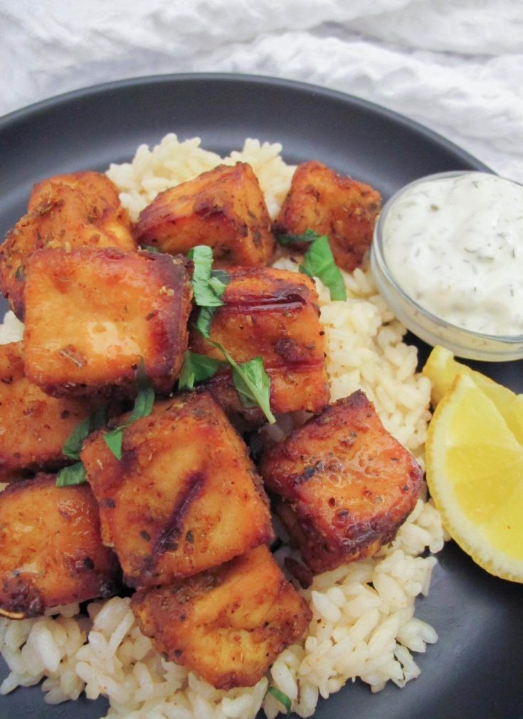 Greek tofu pieces served with rice, tzatziki and two lemon wedges on a black plate.