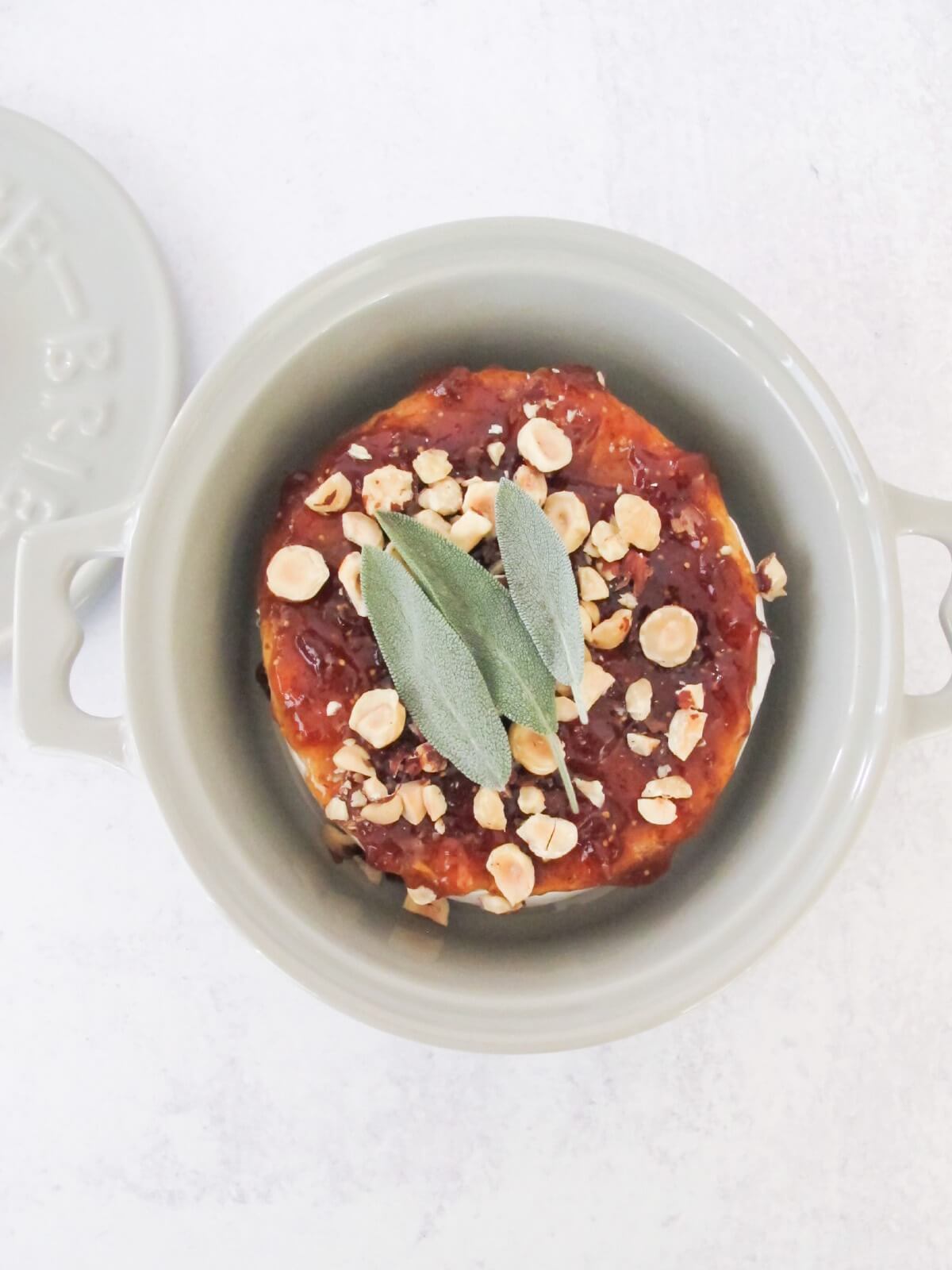 a wheel of brie cheese topped with fig jam, crushed hazelnuts, and fresh sage.