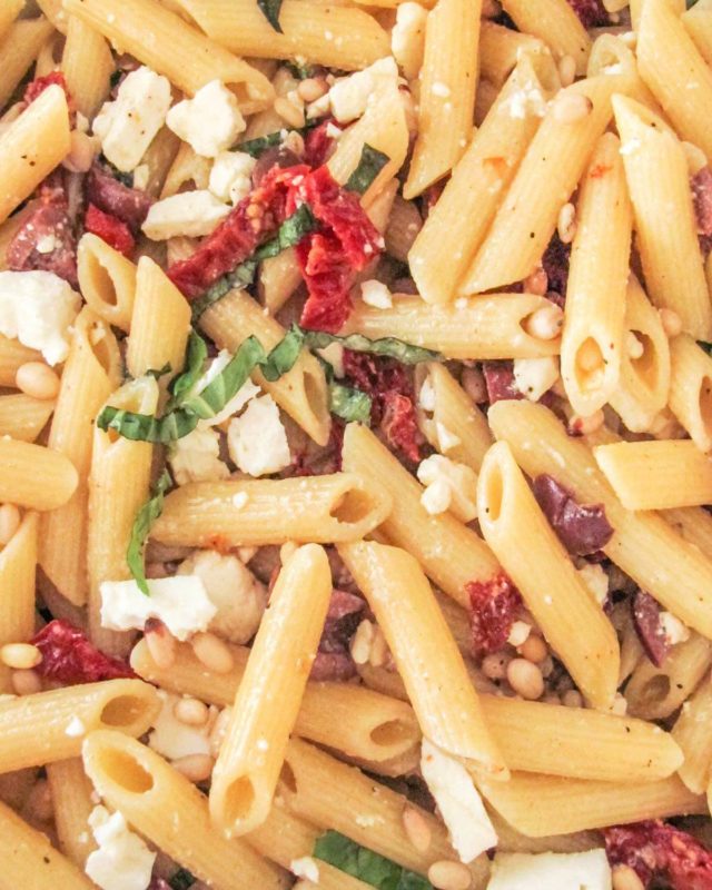 close up photo of greek pasta salad with penne, kalamata olives, sundried tomatoes, basil, pine nuts, and creamy Greek dressing.