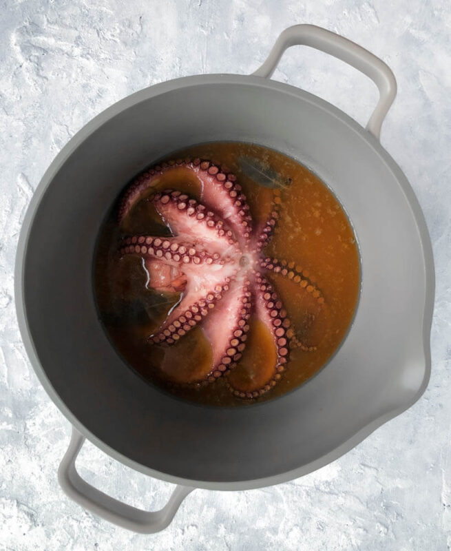 A whole octopus simmering in a large pot with white wine, olive oil, white wine vinegar, bay leaves, and peppercorns.