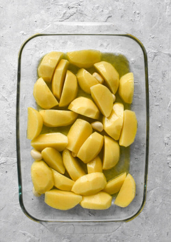 Raw yukon gold potatoes cut into quarters in a baking dish with olive oil and whole peeled garlic cloves.