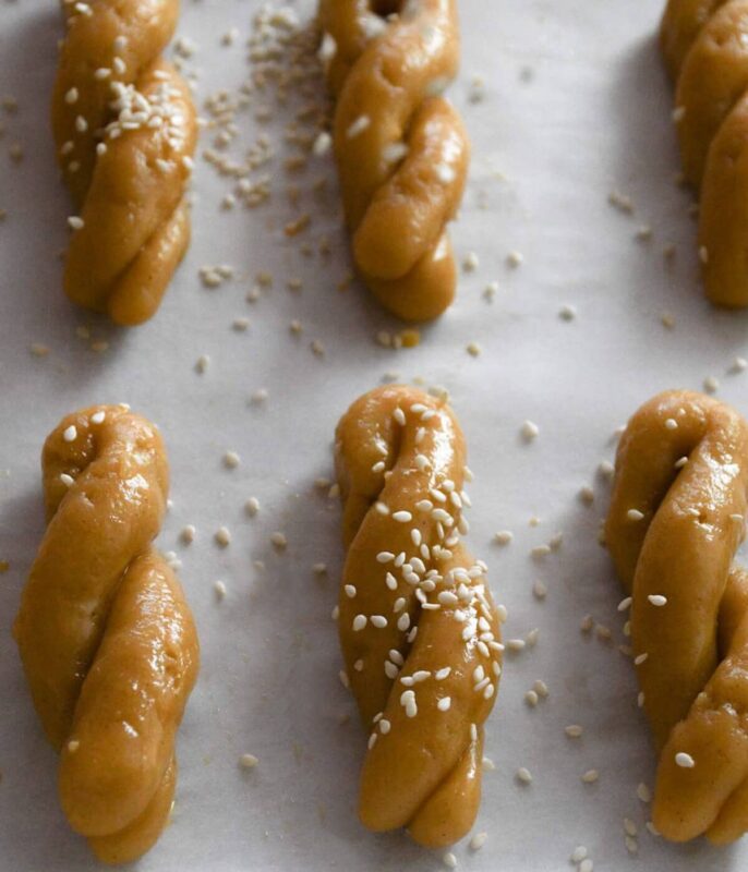 Koulourakia cookies uncooked on a baking sheet sprinkled with sesame seeds.