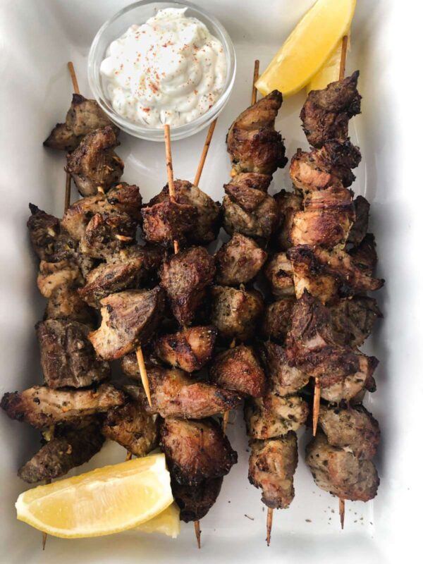 baked pork souvlaki skewers in a white baking dish with a side of tzatziki sauce.