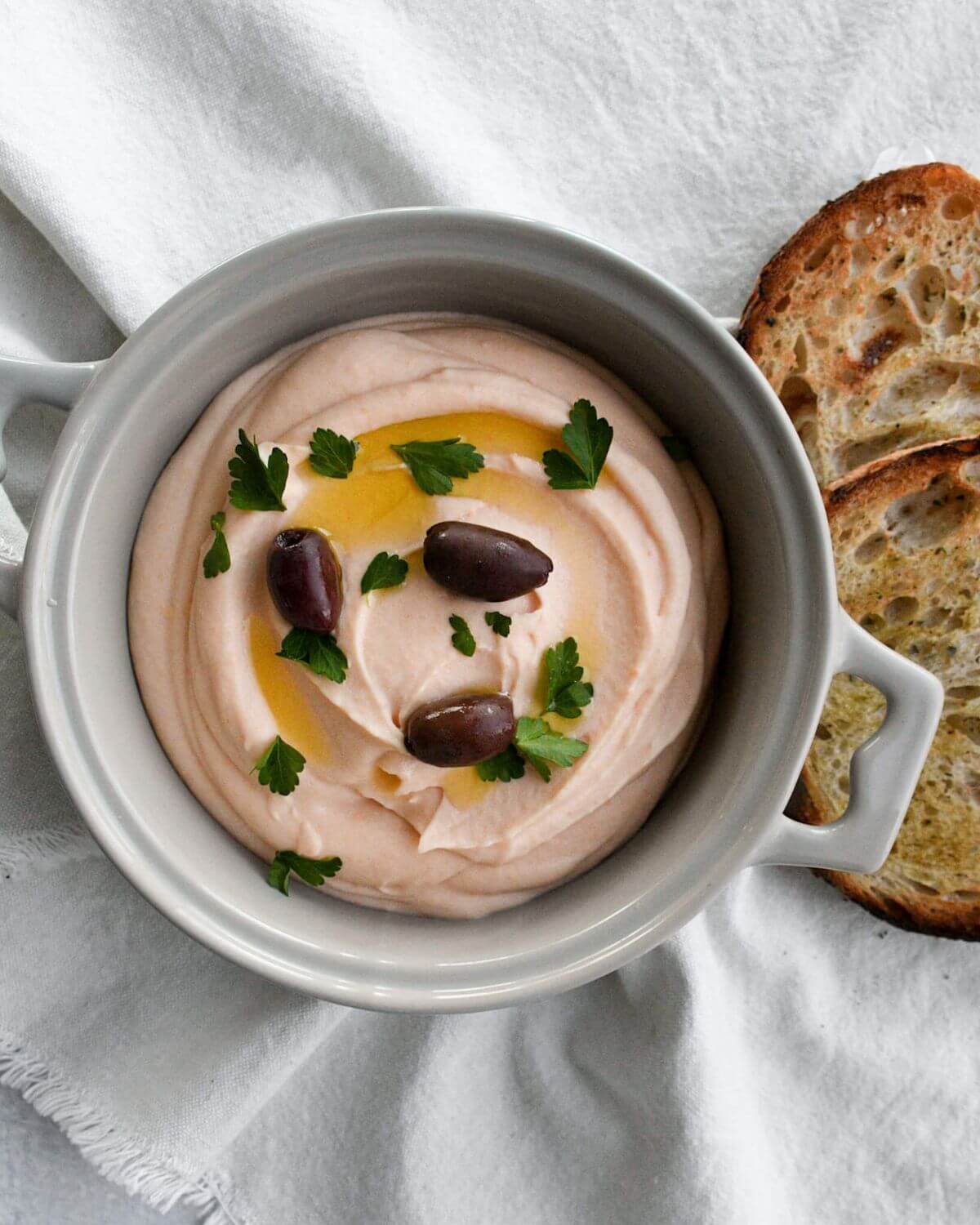 taramasalata dip in a light grey bowl topped with olive oil, parsley, and olives.