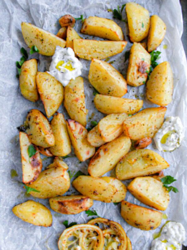 Oven roasted Greek lemon potatoes on white parchment paper with fresh parsley and tzatziki.