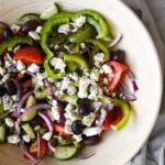 Greek salad tossed in olive oil with crumbled feta, kalamata olives, and dried oregano in a large wooden bowl.