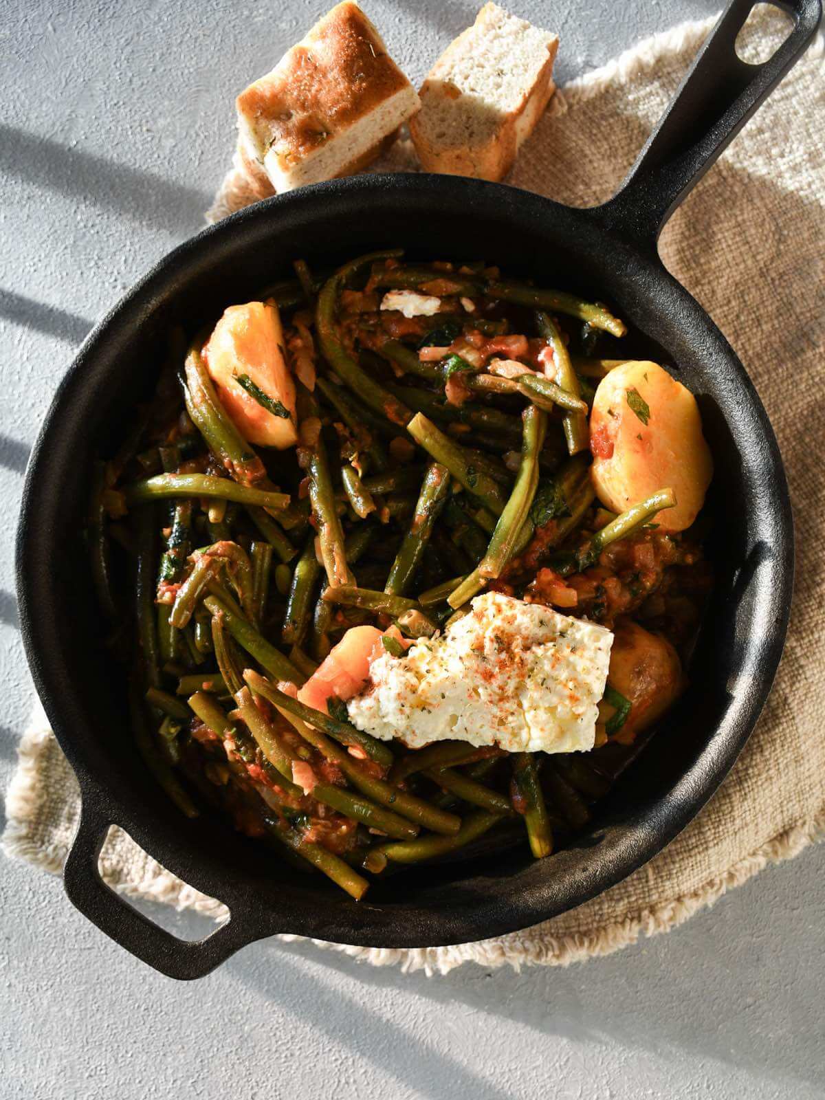 Greek green beans with tomato sauce and potatoes (fasolakia) with feta cheese displayed in a black cast iron skillet.