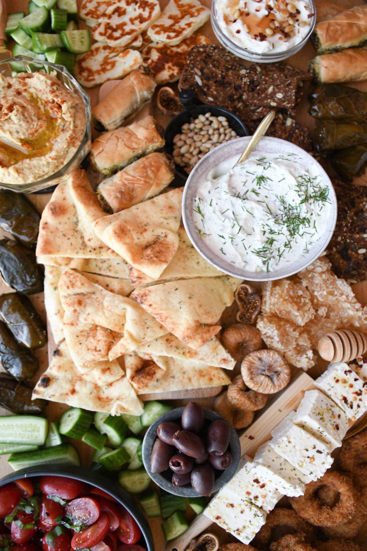 greek appetizer platter with tzatziki, pita bread, spinach pie, honey, dried figs, feta cheese, cucumber, and olives on a wooden board.