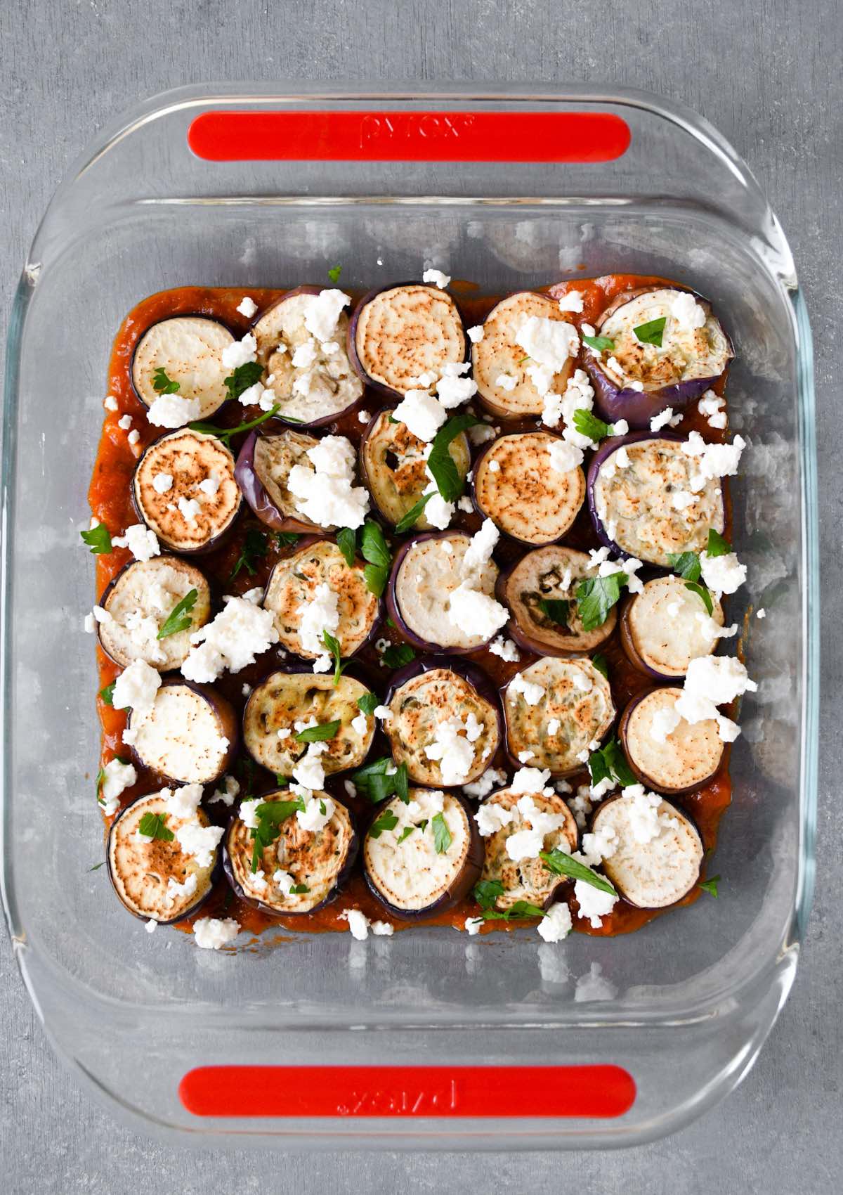 cooked eggplant slices on top of tomato sauce in a baking dish with feta and parsley.