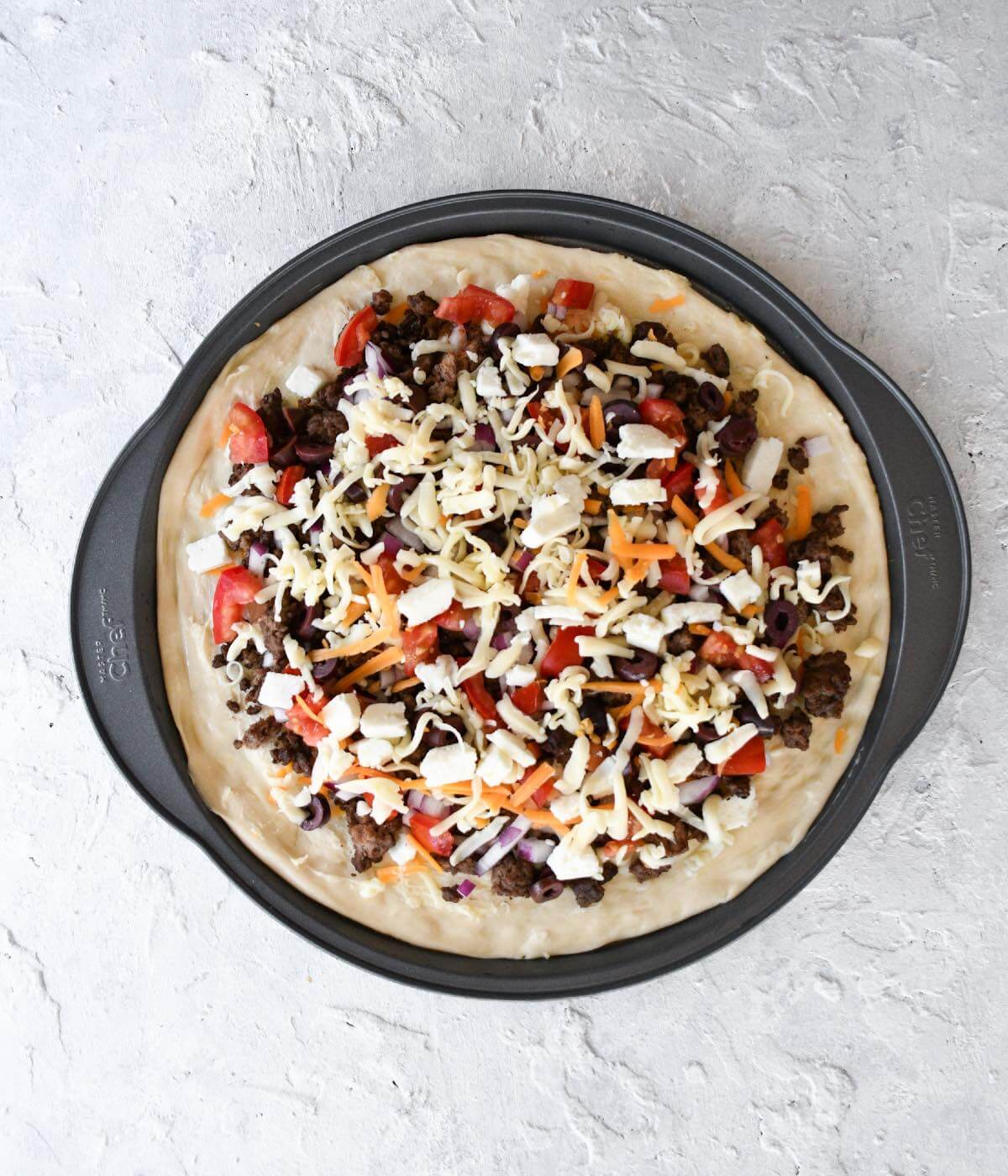 homemade pizza with garlic butter, mixed cheese, cooked ground beef, tomatoes, red onions, and feta in a 12" pizza pan.