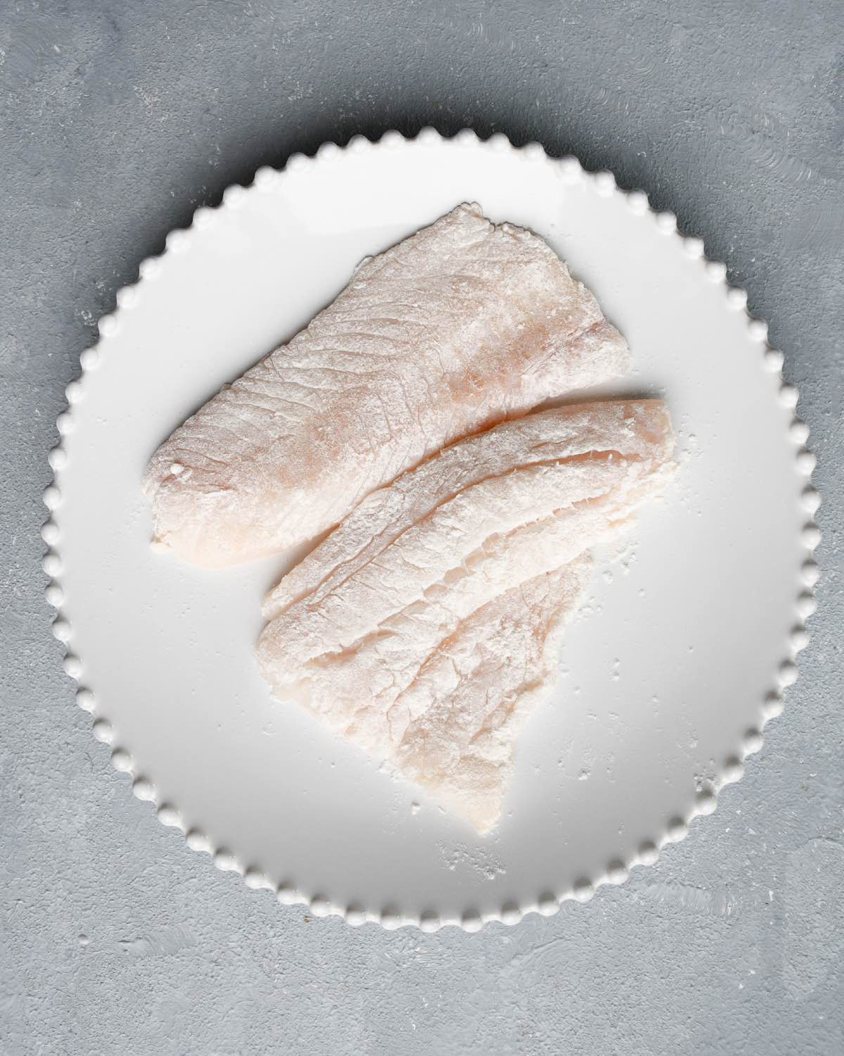 two pieces of floured cod on a white plate.