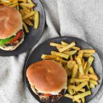 two greek chicken burgers with brioche buns with fries on black round plates.