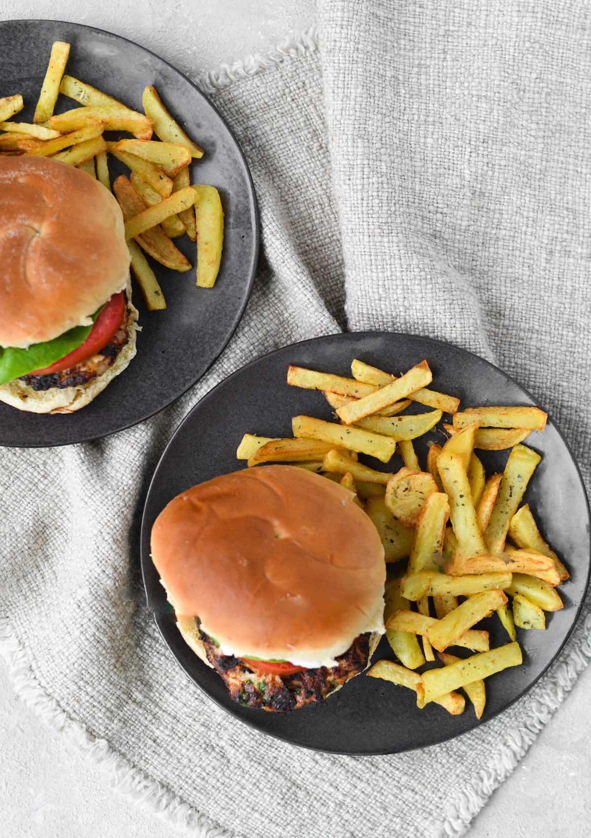 two greek chicken burgers with brioche buns with fries on black round plates.