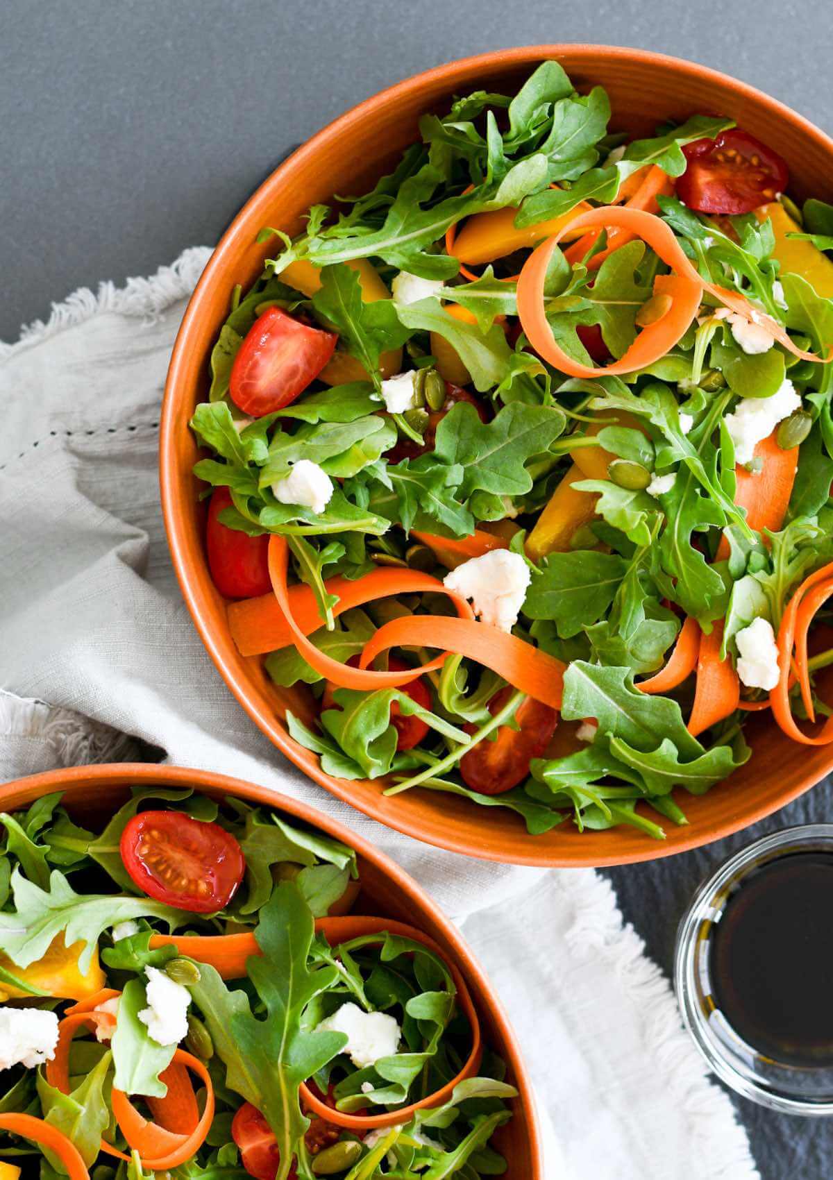 arugula salad in orange round bowls with cherry tomatoes, crumbled goat cheese, ribbon carrots, and bell peppers.