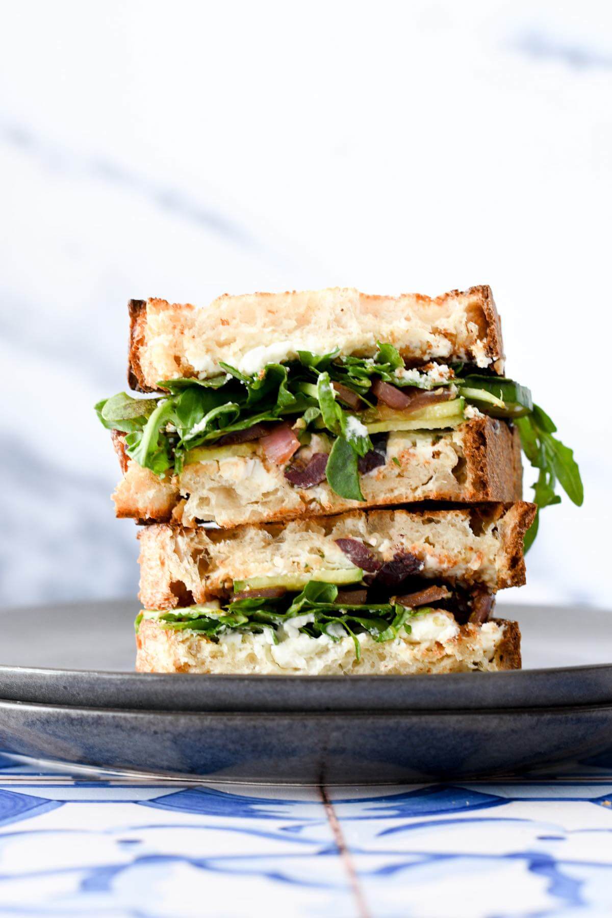 two greek sandwiches on top of each other. Pieces of sourdough bread with cucumber slices, creamy feta cheese, kalamata olives, pickled onions and arugula.